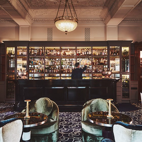 The Coburg Bar. The image shows tables and chairs in the foreground with a well-stocked bar in the background and a bar tender looking at the alcohol bottles.