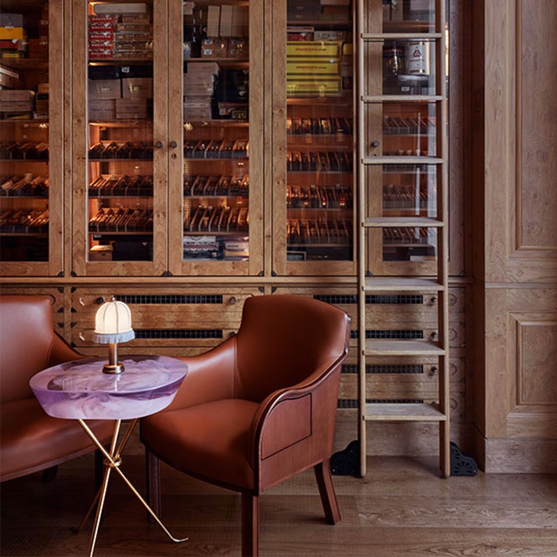 The Connaught Cigar Merchants detail shot of table and armchair in front of the glass doors cupboard that contains the cigar collection.